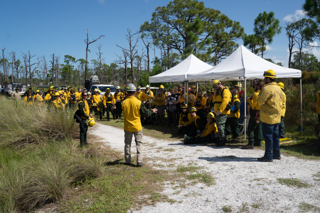 A large group of people are gathered together listening to a speaker. They are outside on a gravel pathway and are all wearing the same fire protective gear and hard hats. 