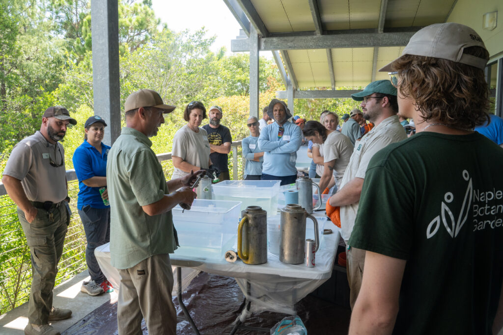 People gather around a table while a person is speaking and showing the group parts of a fire torch. 