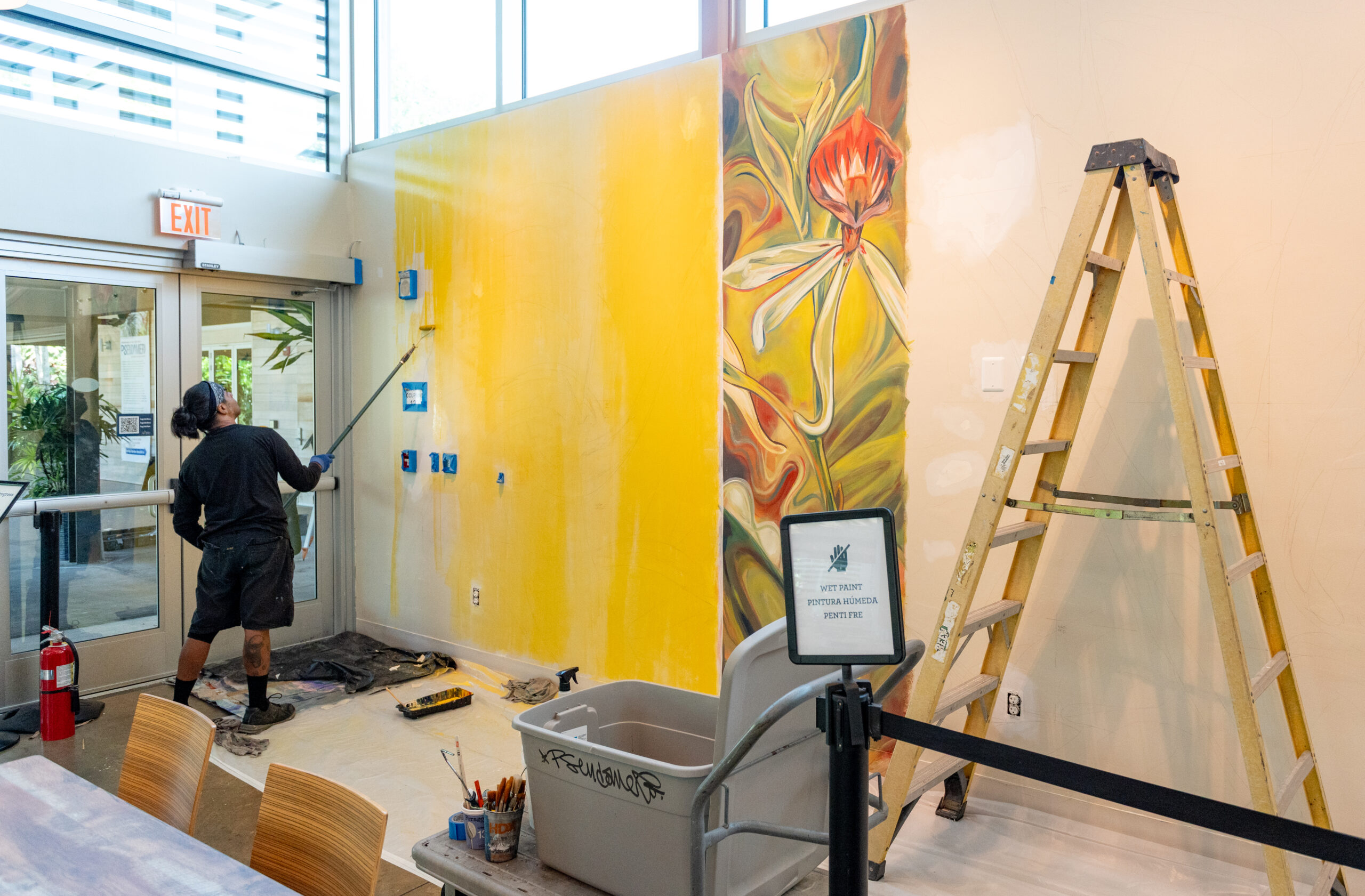 A person paints a wall yellow with a paint roller, a section of the wall next to them is painted with large flowers. There is a ladder to the right.