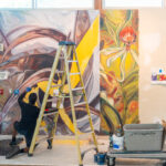 A person works on painting a mural on an indoor wall. There is a ladder and a cart with painting supplies near them.