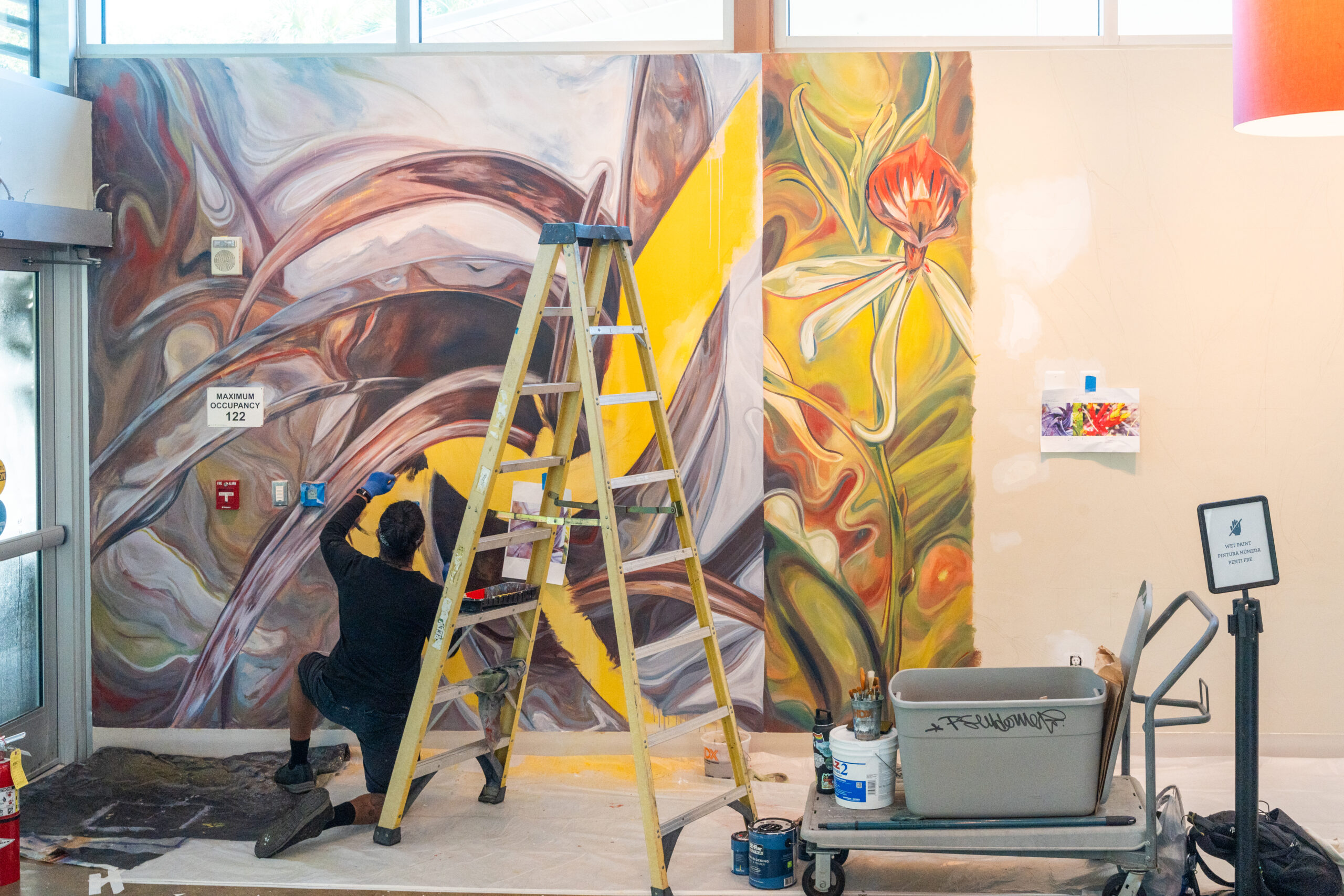 A person works on painting a mural on an indoor wall. There is a ladder and a cart with painting supplies near them.