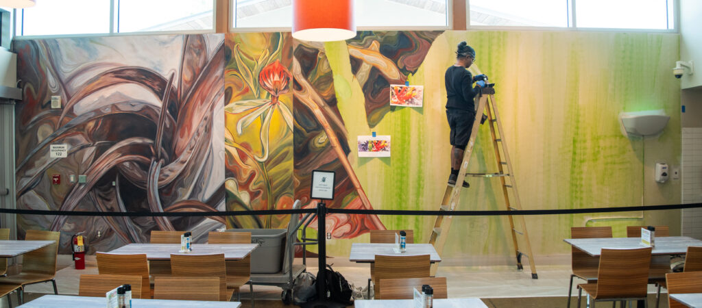 A person stands on a ladder with paint in front of a large mural on a wall in an indoor space near rows of dining tables.