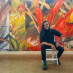 A person poses in front of a mural. On the left there is a macro painting of a tillandsia, next to that an orchid, and on the far right a red bromeliad flower.
