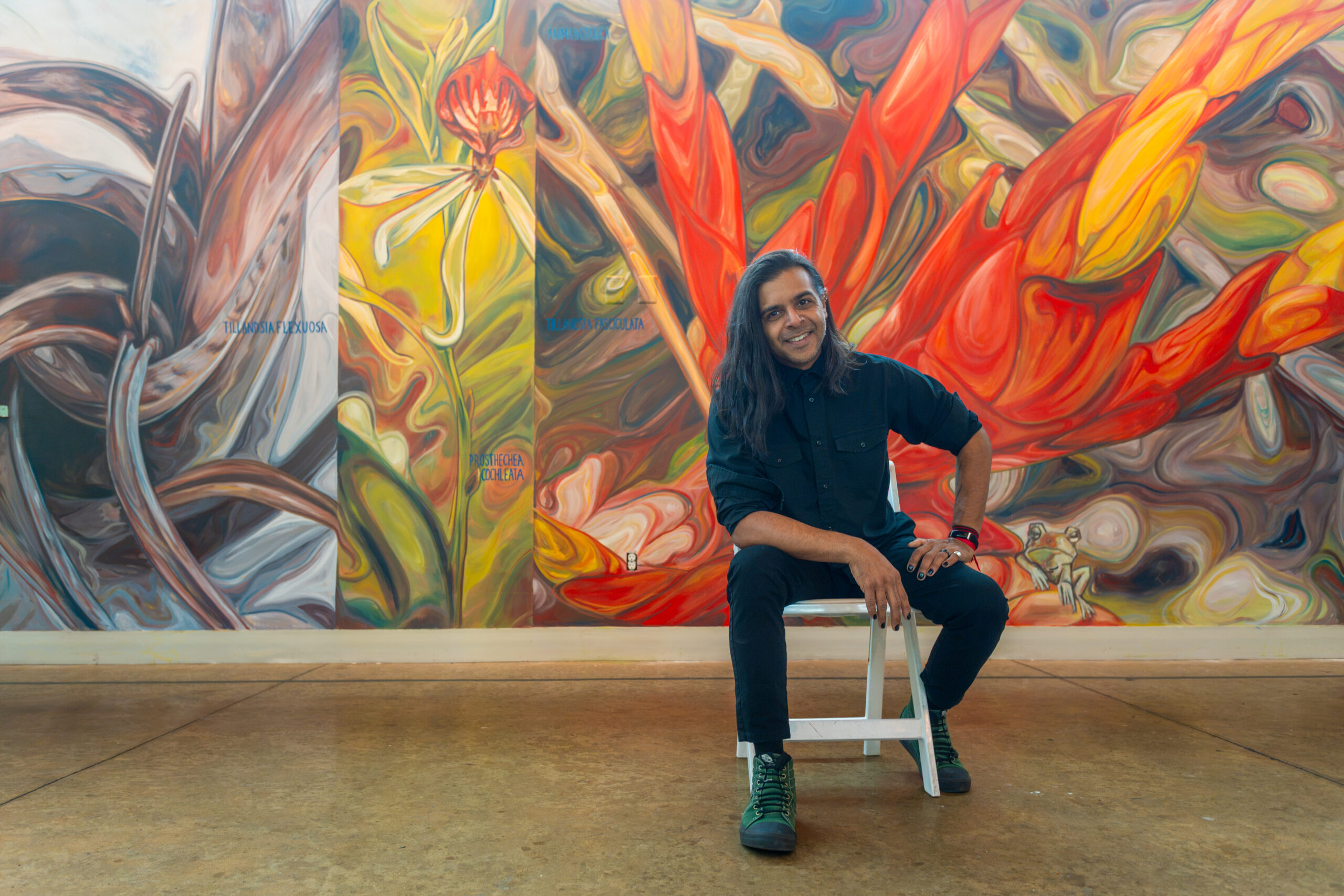 A person poses in front of a mural. On the left there is a macro painting of a tillandsia, next to that an orchid, and on the far right a red bromeliad flower.