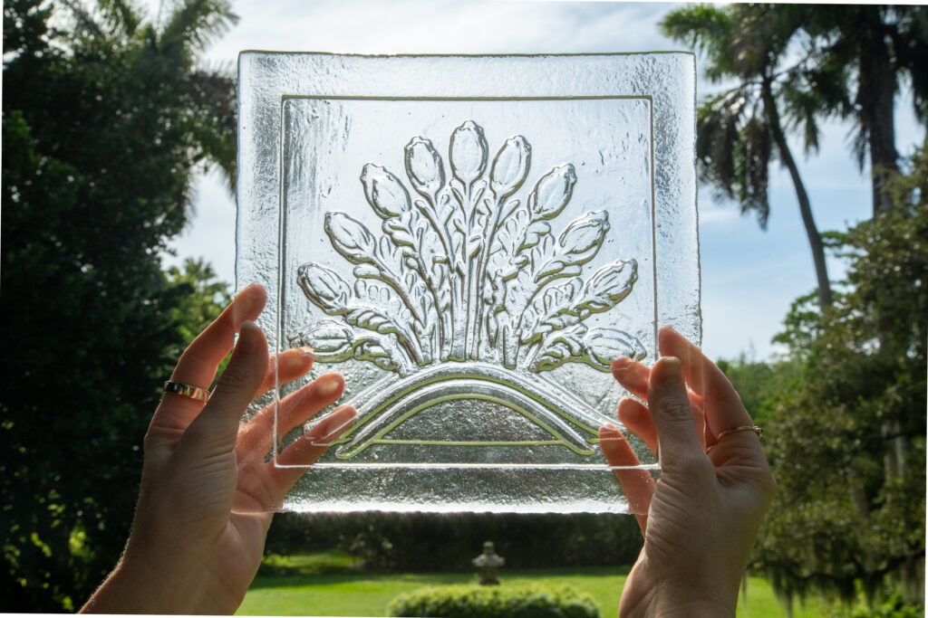 A glass tile with a decorative floral seed design is held up against the sky to see light pass through it
