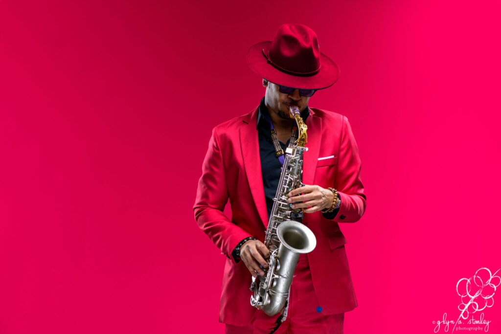 A person wearing a pink suit stands against a pink background while playing a saxophone.