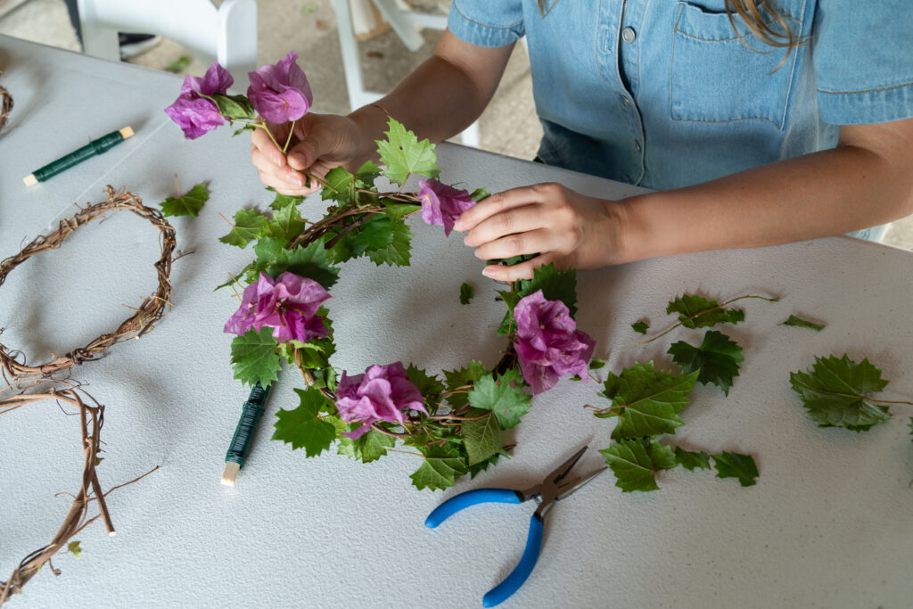 A person sits at a table and adds purple flowers to a woven crown with green leaves embedded in it.