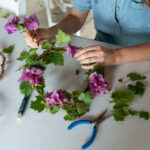 A person sits at a table and adds purple flowers to a woven crown with green leaves embedded in it.