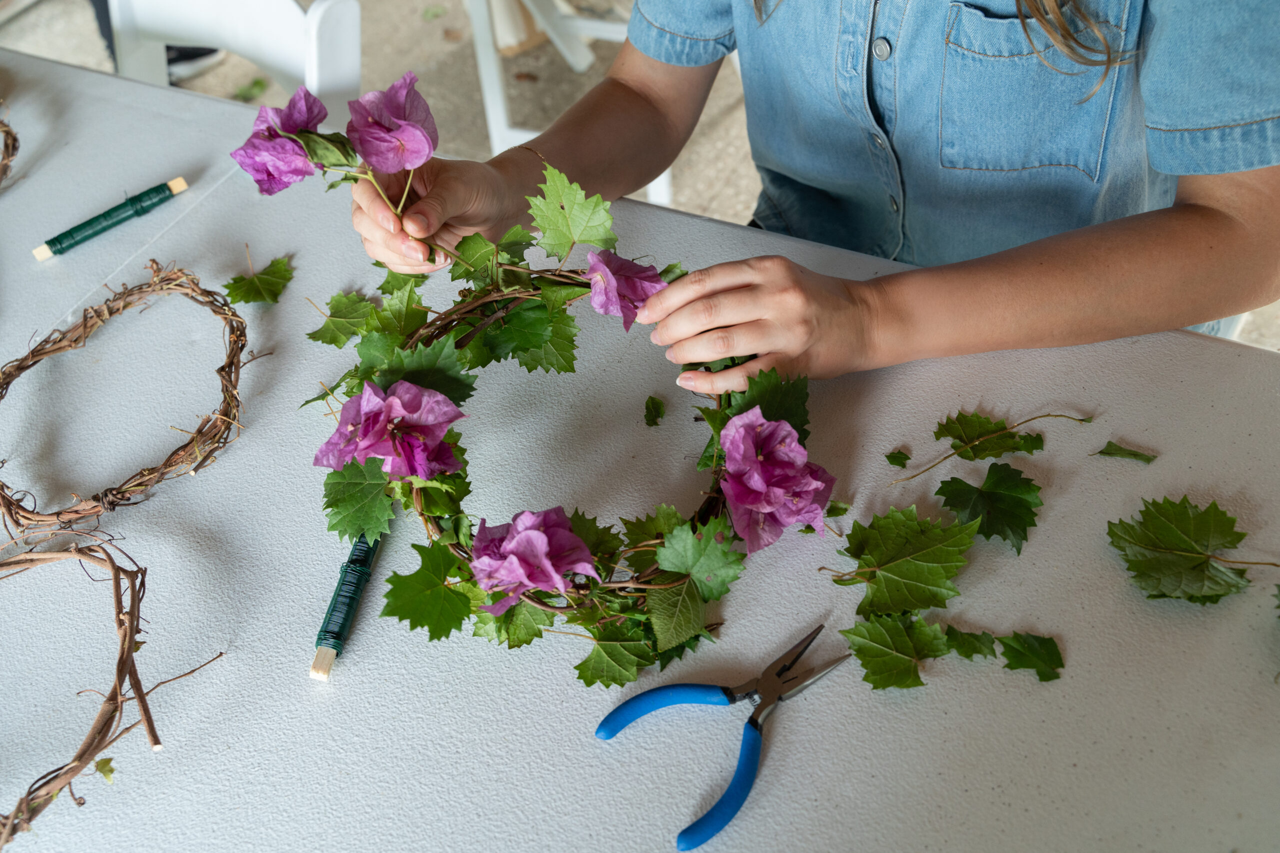 A person sits at a table and adds purple flowers to a woven crown with green leaves embedded in it.