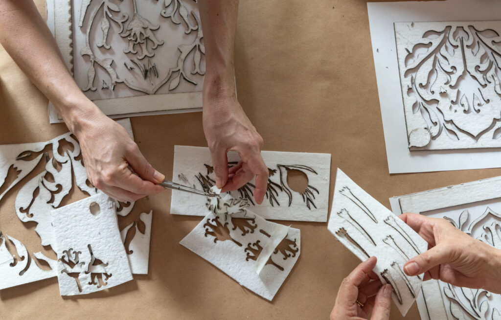 Three hands work to cut out designs of seeds on white paper.
