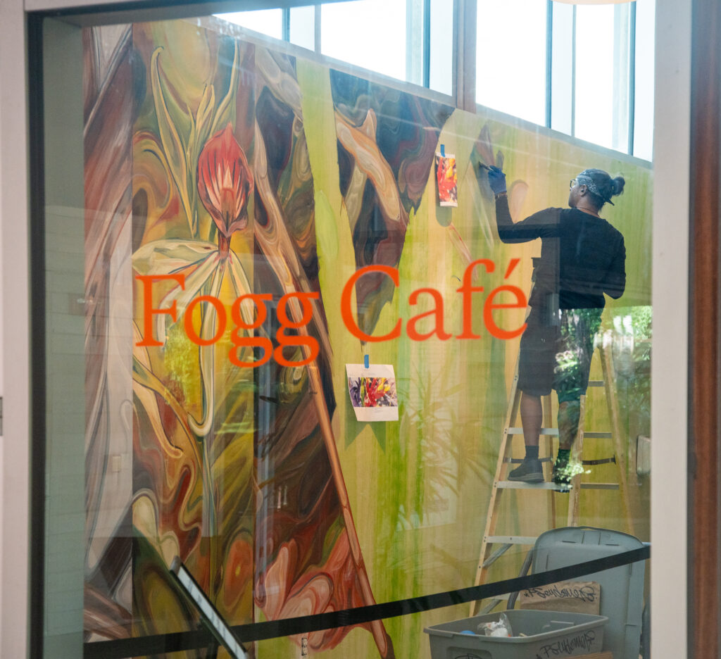 Viewed through a window with a Fogg Café label on the glass, a person stands on a ladder and paints a mural on the wall.