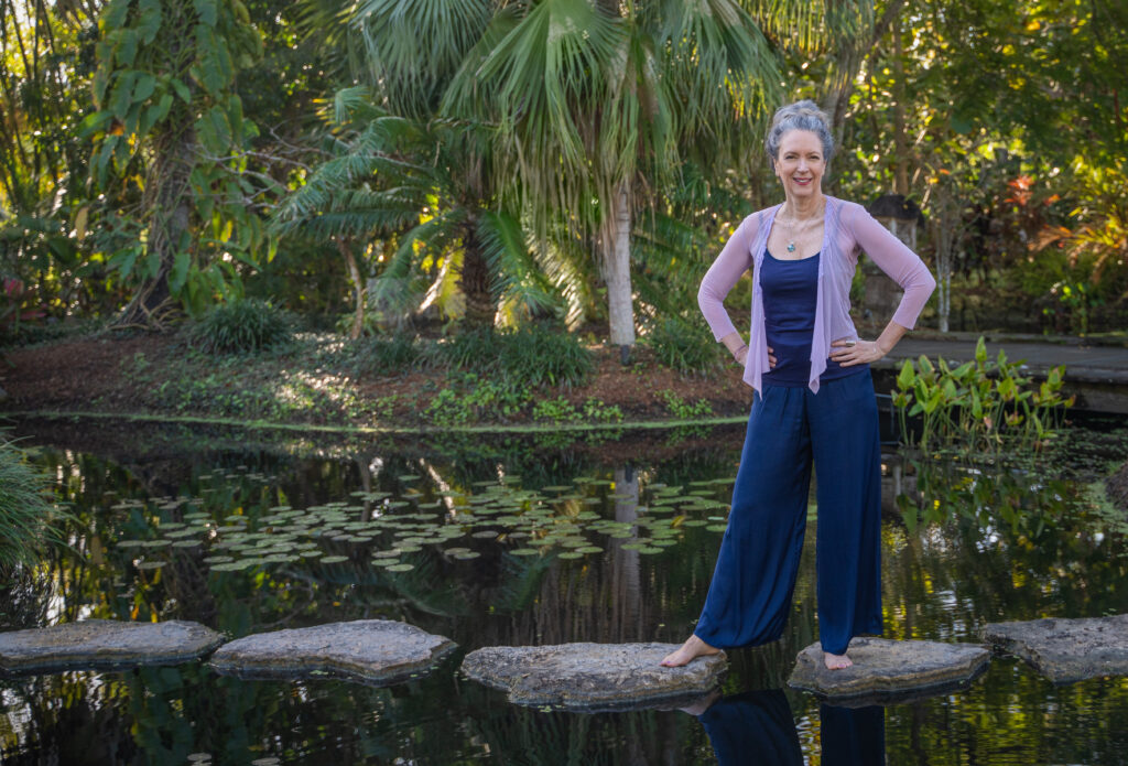 A person poses for a photo while standing on stepping stones laid across a pond. There are waterlilies in the water and palm trees in the background.