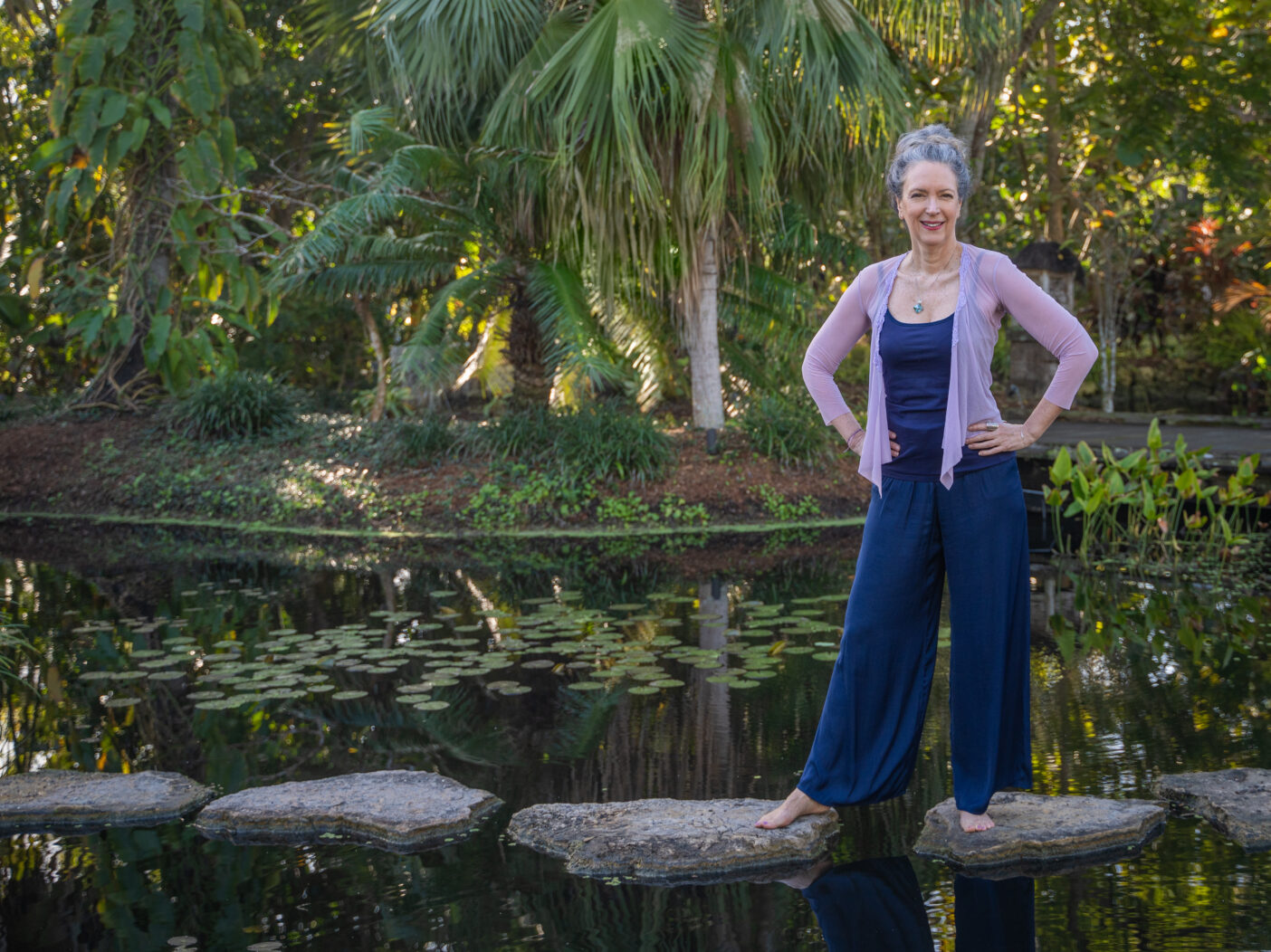 A person poses for a photo while standing on stepping stones laid across a pond. There are waterlilies in the water and palm trees in the background.