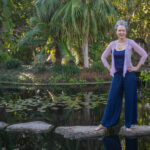 A person poses for a photo while standing on stepping stones laid across a pond. There are waterlilies in the water and palm trees in the background.