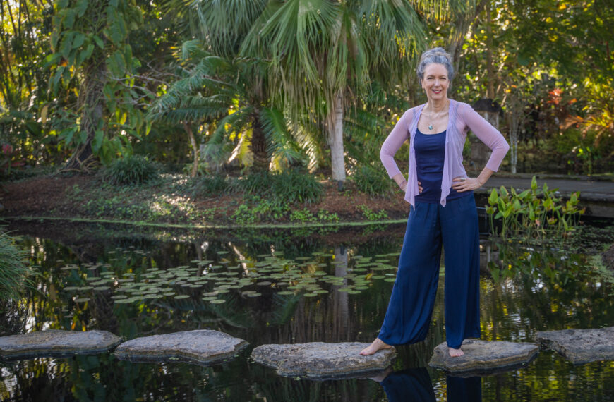 A person poses for a photo while standing on stepping stones laid across a pond. There are waterlilies in the water and palm trees in the background.