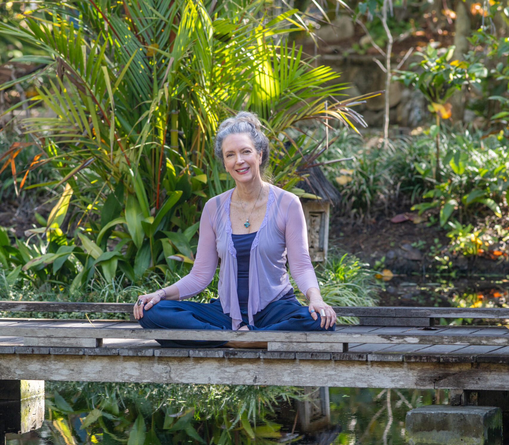 A person sits cross-legged on a boardwalk that spans a pond surrounded by tropical plants.